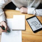 Eine Person in weissem Hemd schreibt mit einem grünen Stift auf ein Blatt Papier, während daneben ein Tablet mit einer einfachen Rechenaufgabe (150 + 250 = 400) auf dem Tisch liegt.